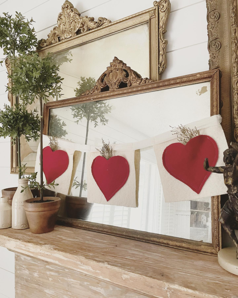 A vintage Valentine garland made up of felt hearts on felt square-shaped pieces of fabric, hanging across a vintage mirror on a vintage fireplace mantel.