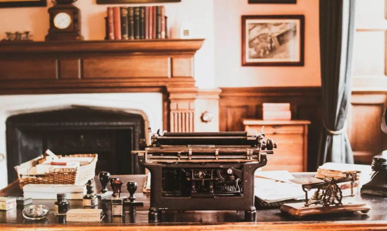 The interior of an old office with vintage office decor items on a wooden desk such as a typewriter, wooden basket, seal stamps, and a fireplace in the background.