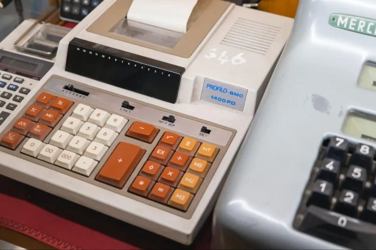 A 1980s calculator with white, orange, and yellow buttons, surrounded by other vintage calculators.