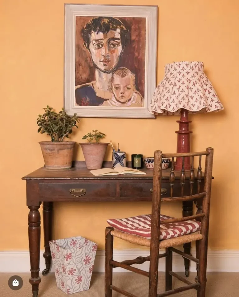 A home office nook with a yellow-orange wall, an antique desk, a vintage wooden chair, a vintage lamp, some vintage accessories, and a painting on the wall.