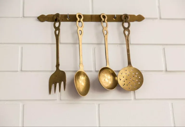 Four vintage brass cooking utensils hanging on a cream-tiled wall.