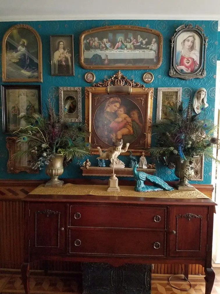 Various religious paintings in ornate antique frames and a wall sculpture of Mary hang on an ornate turquoise wall, with a vintage wooden dresser in front of it, adorned with two bouquets of peacock features, an angel figurine, and a turquoise sculpture of a peacock.