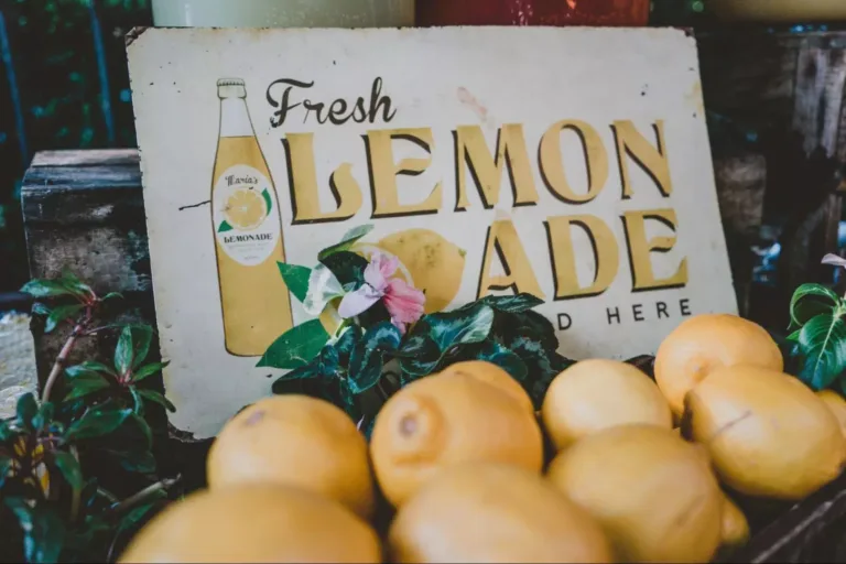 A vintage lemonade sign reading “Fresh Lemonade” in an old-style yellow font, with lemons and plants in front of it.