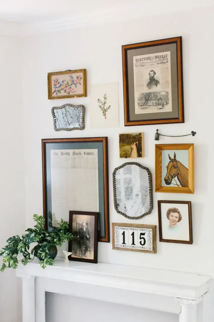 A small gallery wall made up of two old photos, two vintage mirrors, two old newspaper pages, and illustration of a horse, a botanical drawing, and a framed embroidery of birds hanging on a white wall above a white mantelpiece.