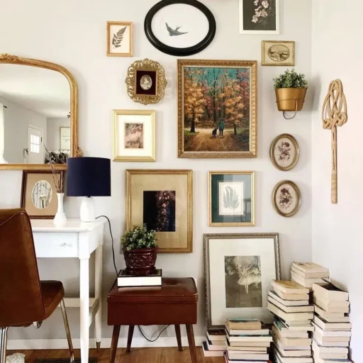 A white wall filled with old drawings and illustrations in oval and rectangular ornate frames; various books piled up in front of it.