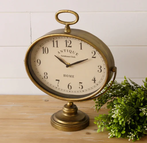 An antique farmhouse table clock on a wooden kitchen countertop with a fake green plant next to it.