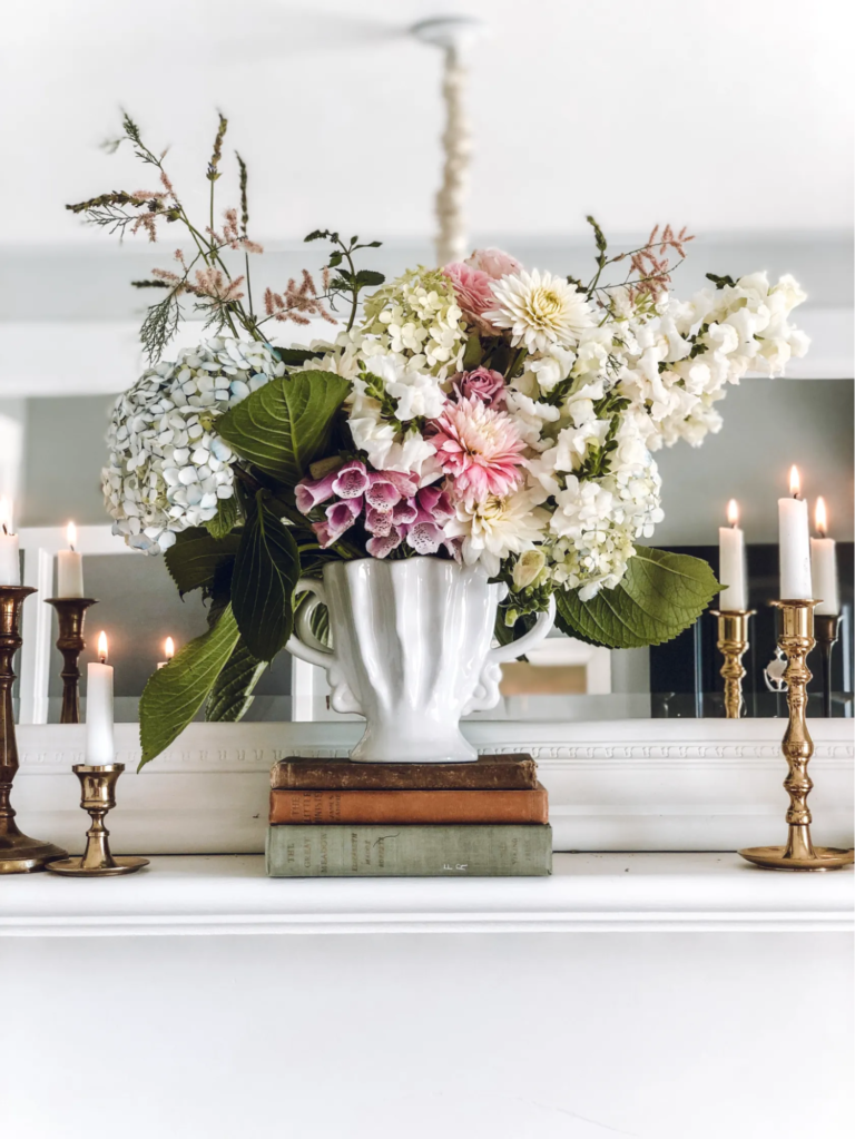 A white vintage mantelpiece with a mirror, pink flowers in a white vintage vase sitting on top of some vintage books, and three white candlesticks in brass candlestick holders.