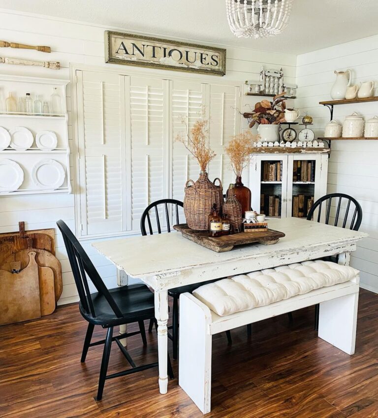 A vintage farmhouse kitchen with a rustic white table, cabinet with white plates, wicker containers and a vintage farmhouse sign reading Antiques.