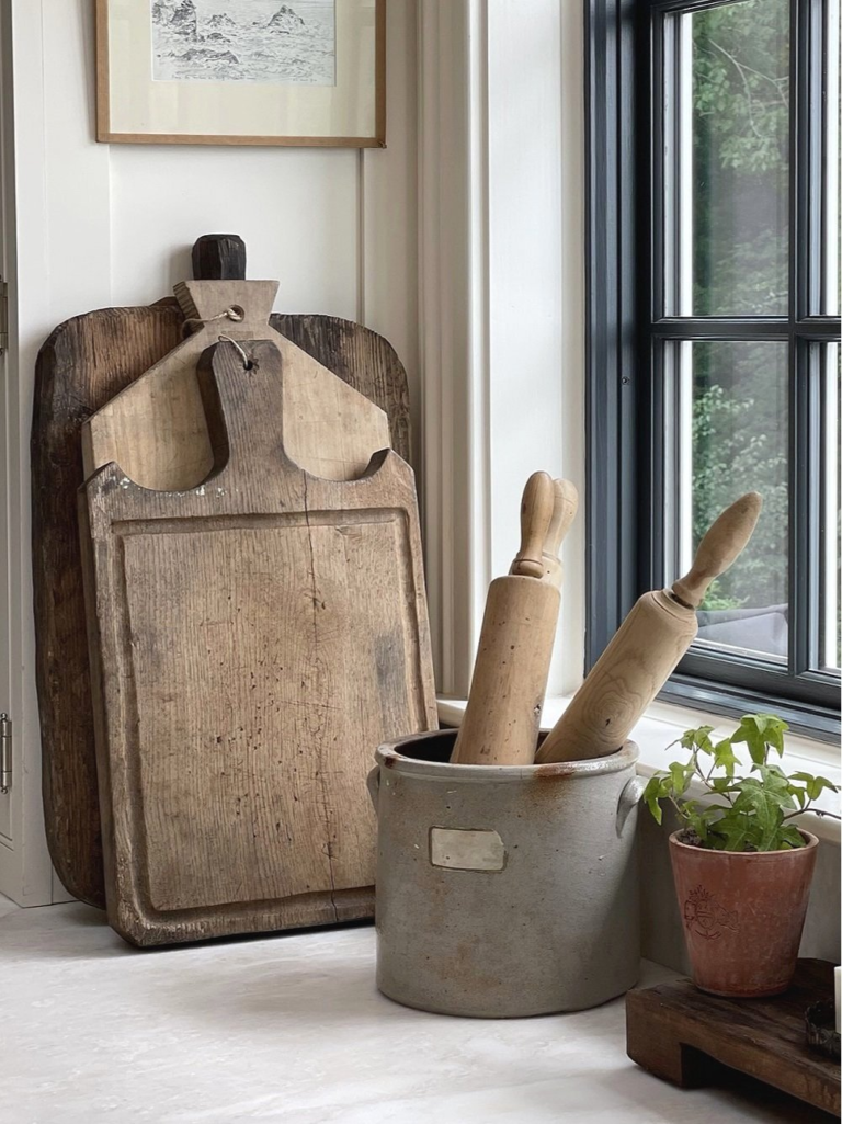 Vintage wooden French bread boards stacked against the wall, a vintage clay container with wooden French rolling pins, and a vintage flower pot with an English ivy plant.