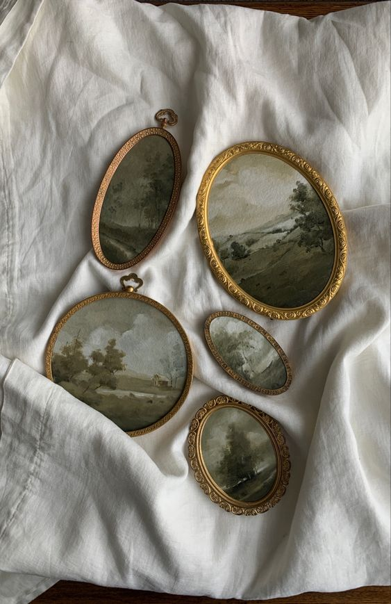 Five antique, gold-tone decorative oval frames with pastoral scene paintings, set on a white linen cloth.