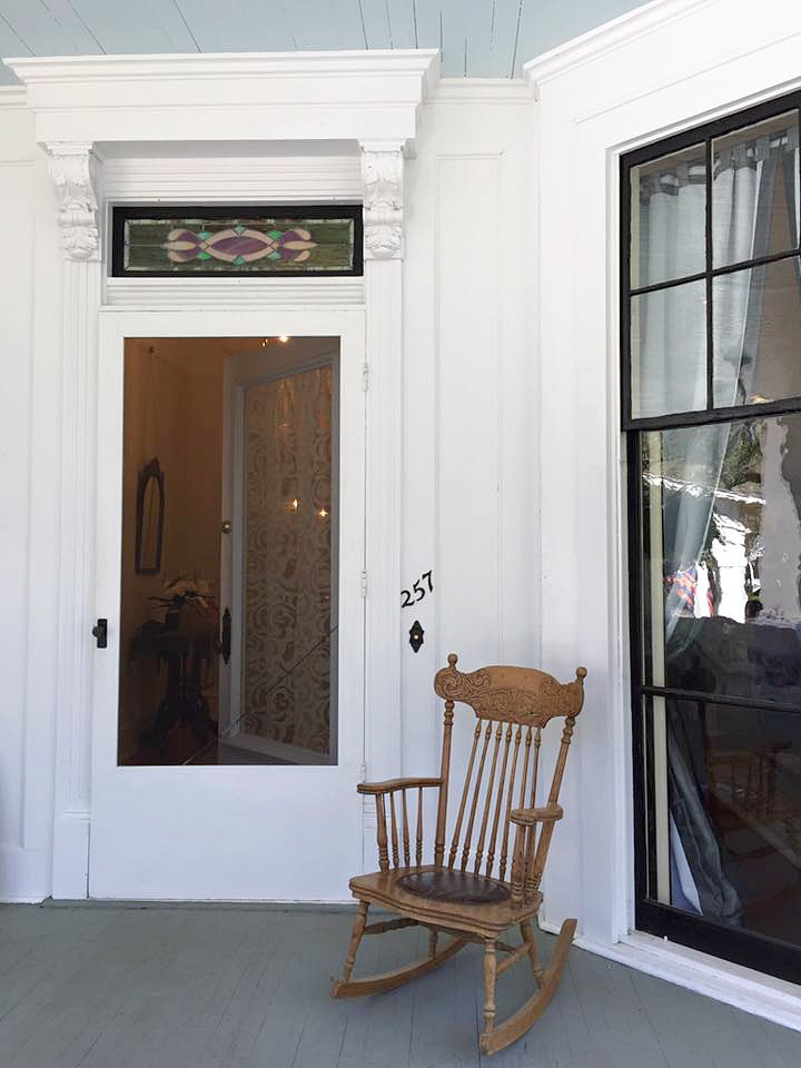 An antique wooden rocking chair on the wooden floor of a Victorian porch with a white ornate door behind it.