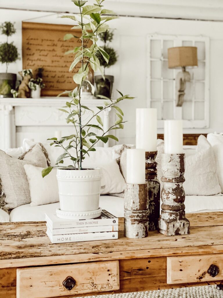 A distressed, light wood rustic coffee table with drawers with white coffee table books and a plant on top and rustic candle holders with white candles. A white vintage mantel in the background.