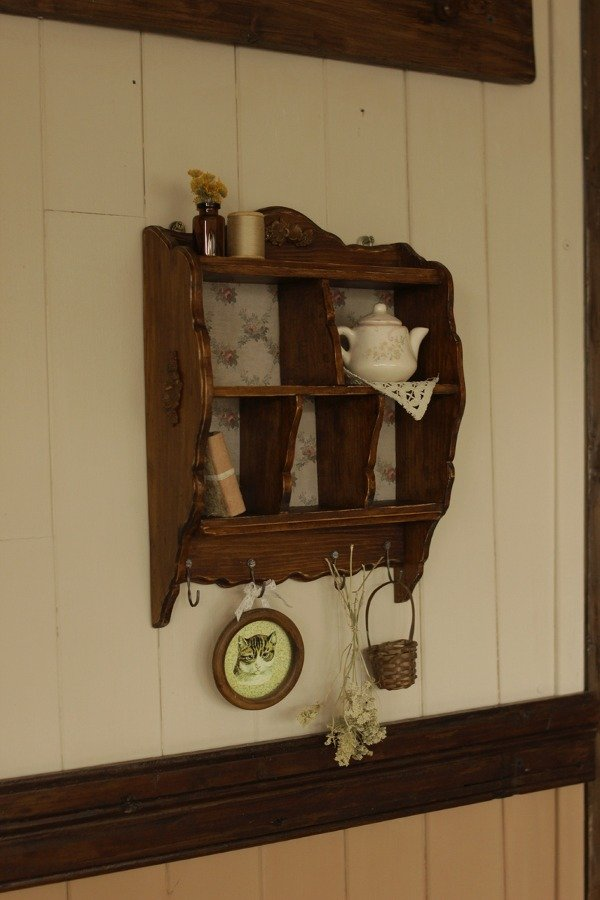 A dark wood vintage display cabinet with a floral vintage wallpaper a vintage teapot, a flower vase, dried flowers, and an oval picture of a cat, hanging on an antique shiplap wall.