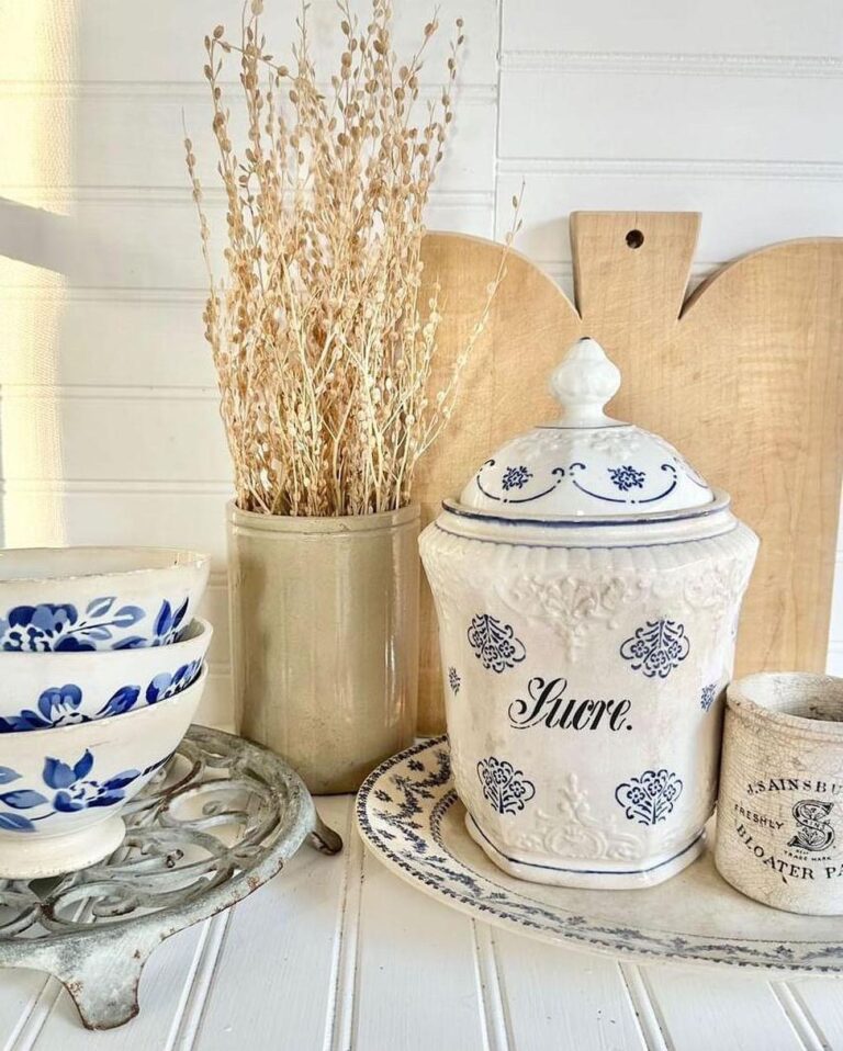 Blue and white vintage farmhouse ceramic containers with dried plants and a wooden cutting board on a white board surface.