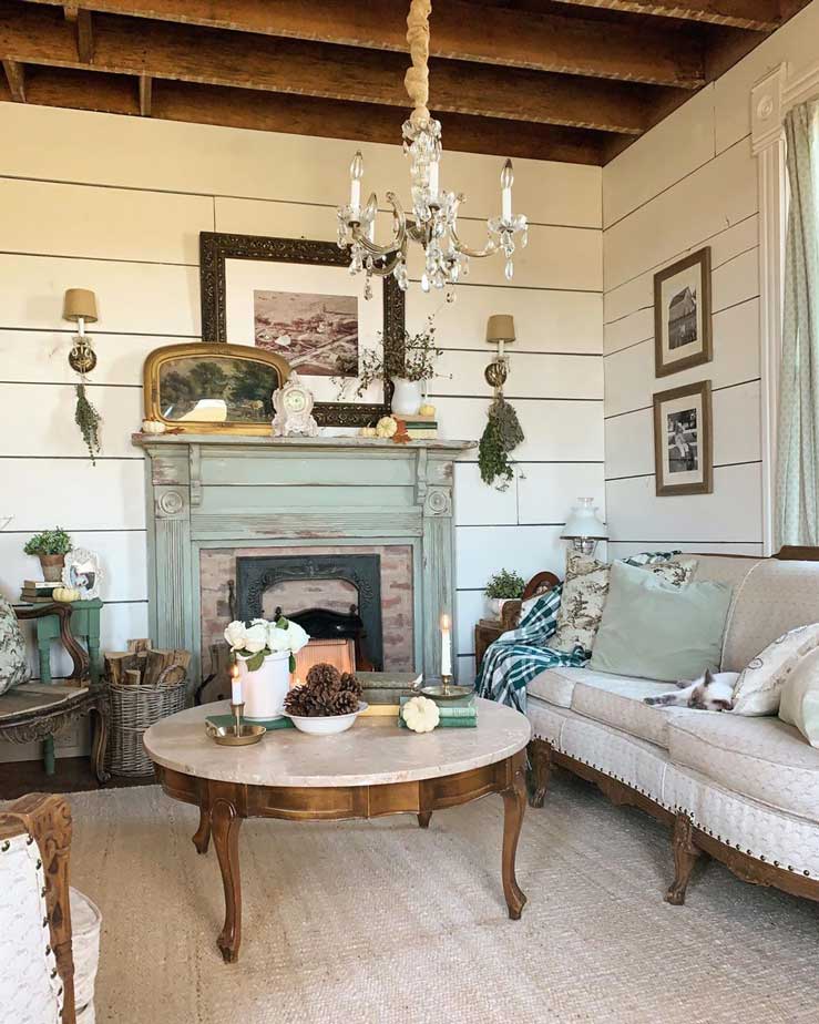 A vintage farmhouse interior with a rustic mantelpiece, white board walls, a vintage couch, round vintage table, wood beam ceiling, vintage pictures on the wall, vintage wall sconces, and a vintage chandelier.