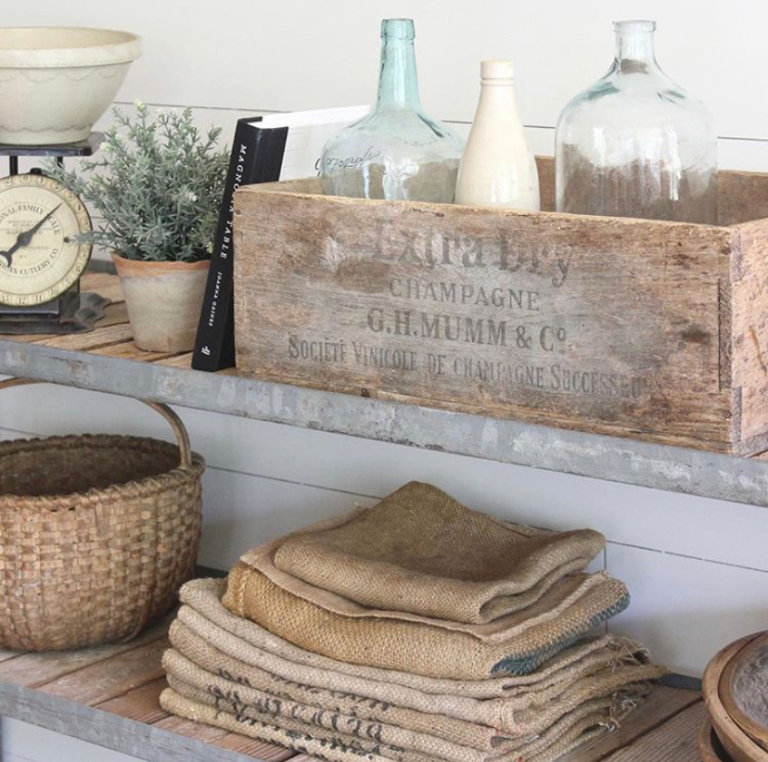 Vintage farmhouse decor with a rustic and distressed wooden crate with an old imprint and vintage glass bottles inside, a stack of organic jute linens, a wicker basket, an old kitchen scale, and a flower pot with French lavender.