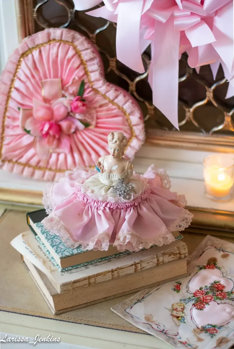 A pink vintage Valentine setup featuring a pink satin ribbon heart-shaped candy box in the background and a small porcelain ballet dancer doll in a pink dress sitting on vintage books.