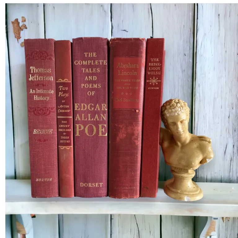 Five vintage books bound in red covers with gold lettering, and a gold mini sculpture of Julius Caesar next to them, all sitting on a shelf with wooden white wall behind.