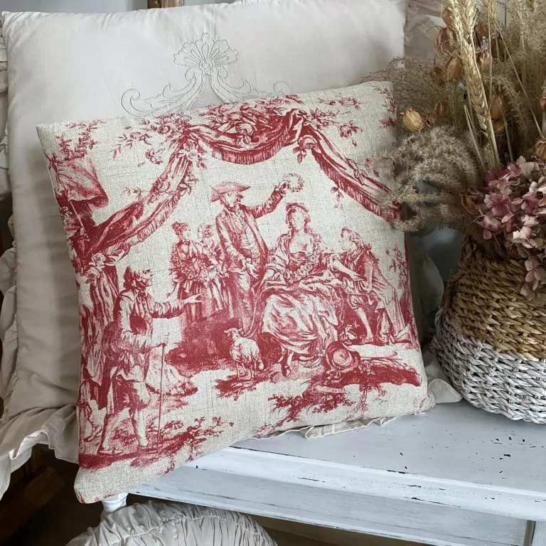 A vintage red and white French toile pillow featuring a pastoral scene, with a wicker basket of dried flowers next to it, sitting on a distressed white shabby bench.