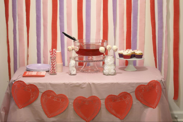 A vintage Valentine’s Day party table with punch, a heart garland, pink paper cups, purple paper plates, and cupcakes on a cake stand. Purple, pink, and red streamers in the background.