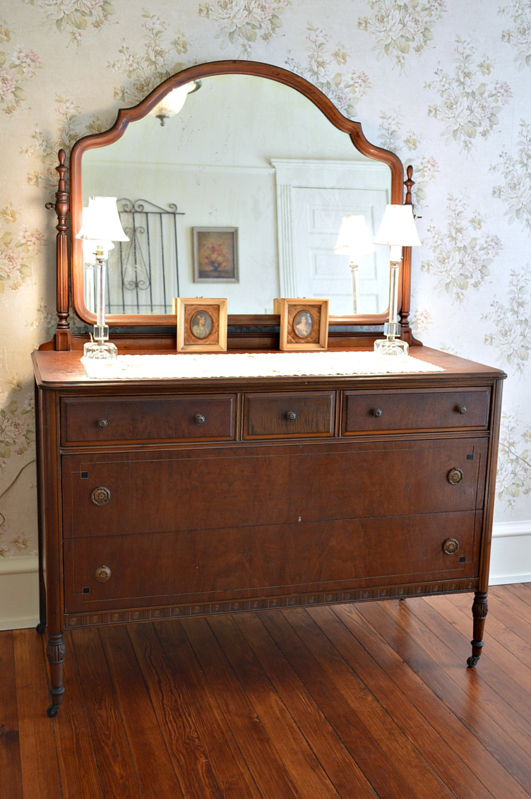 A vintage vanity dresser with a mirror and two lamps with a vintage floral wallpaper in the background.