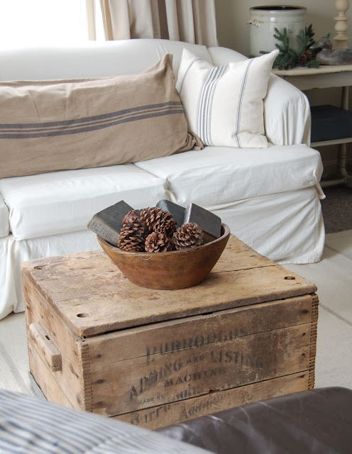 Old wooden crate used as a coffee table, an antique-style wicker chair and a French country house-style couch with pinstripe pillows.