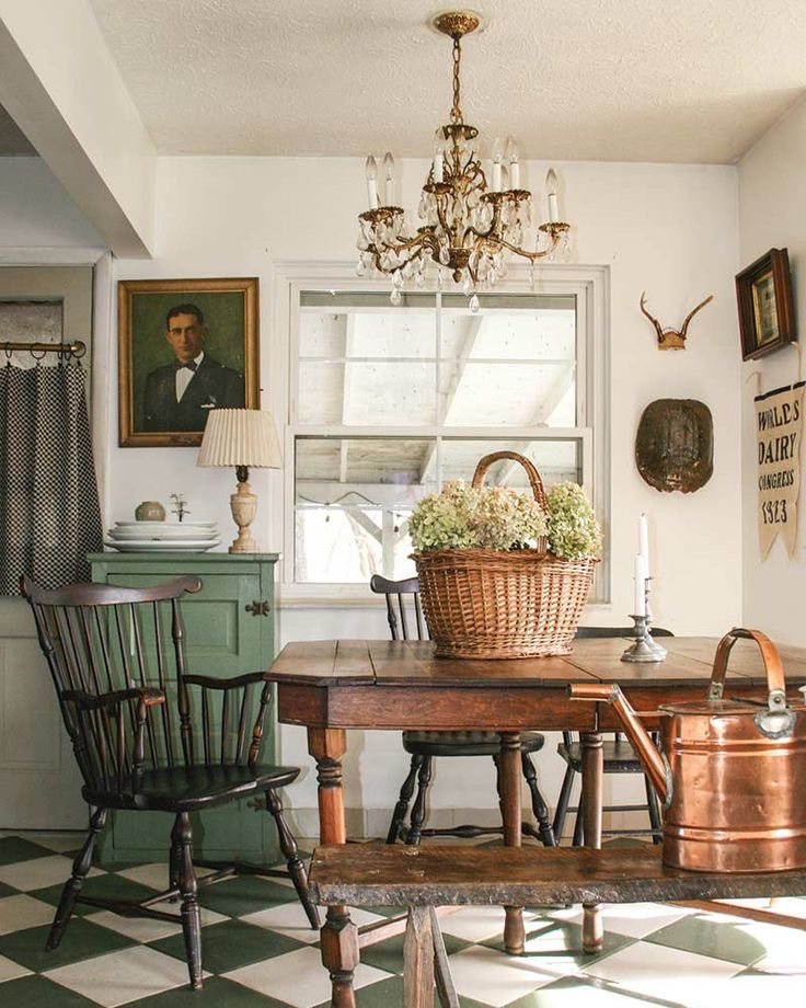 A room with a white and dark green harlequin floor, wooden vintage table with a wicker basket with dried flowers, dark wood hoop-back farmhouse chairs, a sage green vintage cabinet, an antique portrait, a vintage chandelier, and a dark wooden bench with a copper watering can on top.