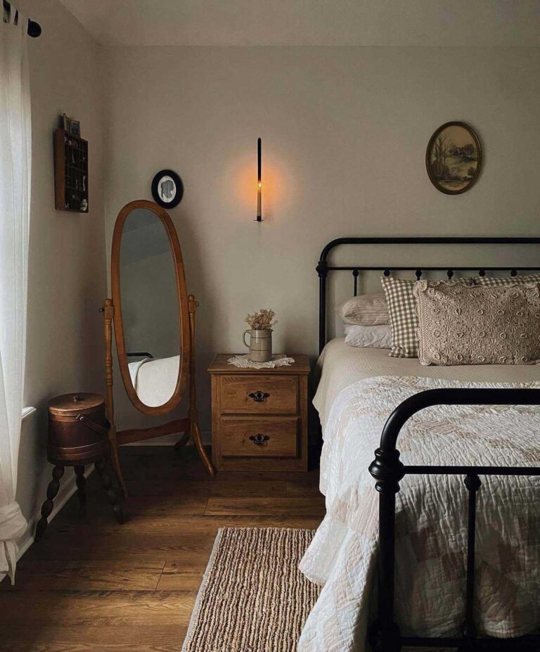 A partially visible bed with a black iron bed frame and vintage bedding made up of a cream-colored quilt and neutral textured pillows. Next to the bed a vintage wooden night stand, a wooden oval cheval mirror, and a jute rug on a wooden floor.