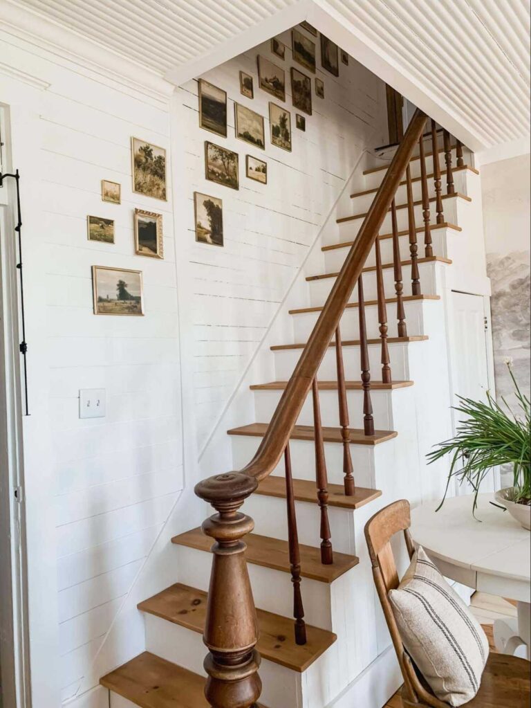 Vintage wooden stairs made of white shiplap and oak boards with a vintage oak newel post, next to a white shiplap wall with a wall gallery made up of vintage frames with antique paintings and illustrations of pastoral scenes.