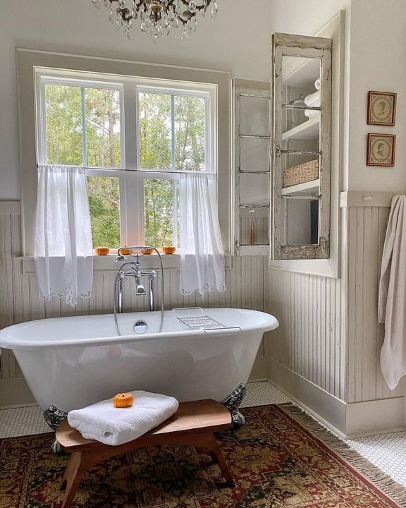 The interior of a vintage farmhouse bathroom featuring an antique-style white clawfoot bathtub, a brown wooden stool, vintage area rug, penny tiles, cream-colored wainscoting walls, a rustic bathroom hutch, and a window with cafe curtains.