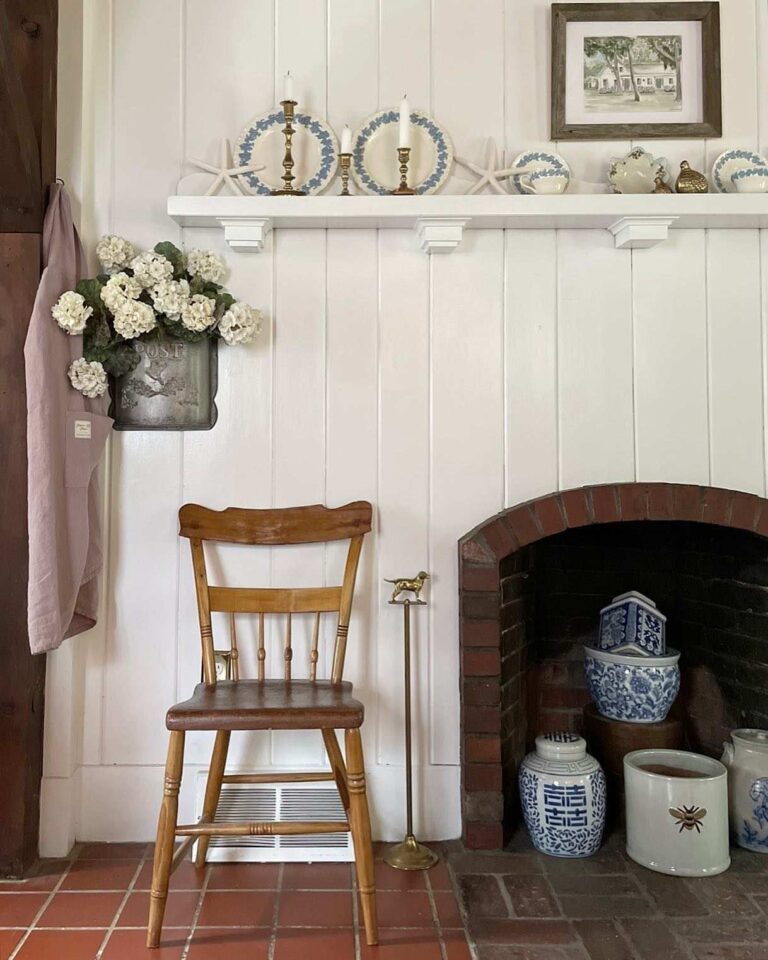 The view of an old brick fireplace inside a white shiplap wall; a white shelf with vintage white and blue plates and brass candlestick holders hangs above the fireplace, and a wooden vintage farmhouse chair stands to the left.