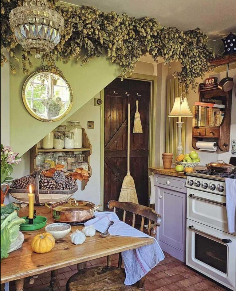 A colorful vintage farmhouse kitchen with light green walls, a wooden table with a vintage wooden chair, an antique gold-frames oval mirror on the wall, a copper dish, small pumpkins, candle and other items on the table, an antique glass hanging lamp, and dried hops as decoration on the top of the wall.