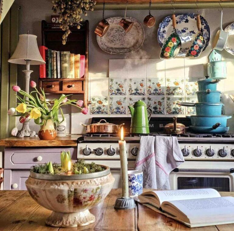 An antique decorative flower pot with hyacinth bulbs, a candlestick, an open book. And a vintage blue and white teacup sit on a vintage wooden table with a vintage colorful farmhouse kitchen seen in the background.