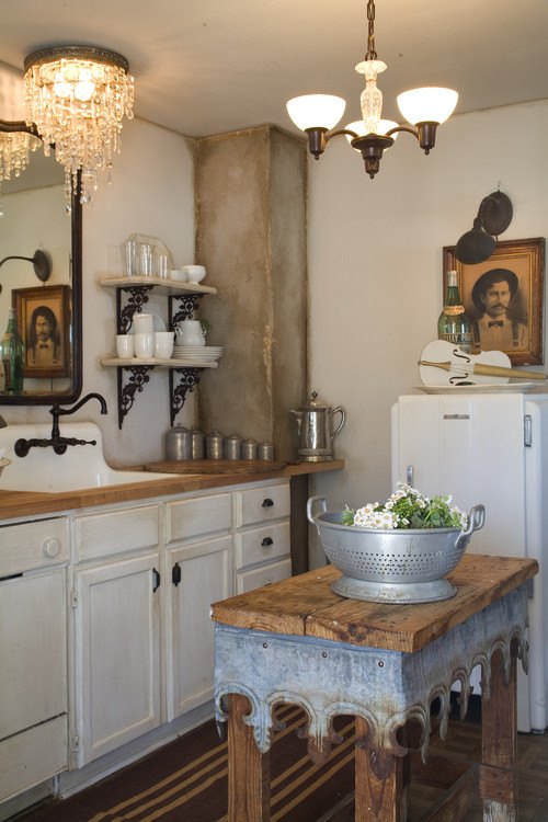 A rustic white vintage farmhouse kitchen with a farmhouse sink, two old portraits, and a reclaimed wood and galvanized metal table used as a kitchen island.