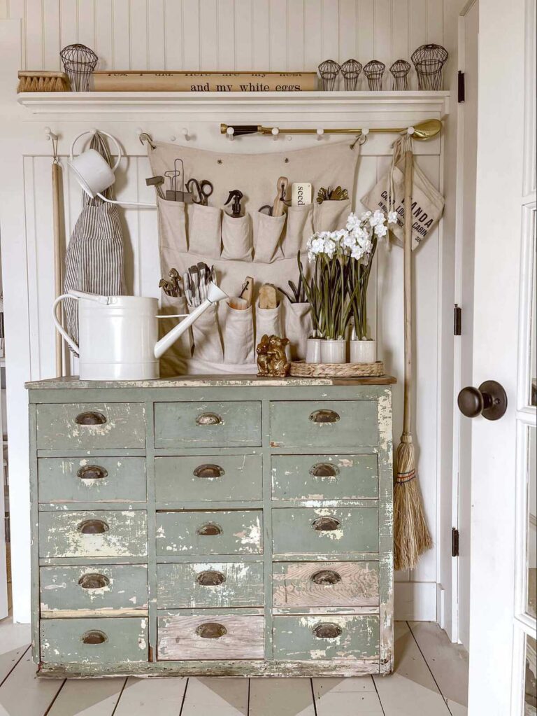 A repurposed wooden apothecary in sage green with a white watering can on top stands against a white shiplap wall. Farm implements hang on the wall in the background.