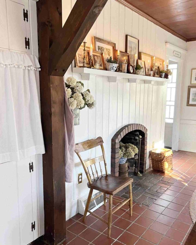 A vintage farmhouse interior with a stone tile floor, stone fireplace, vertical rustic reclaimed wood beam, shiplap wall, white floating shelf with vintage frames, brass candlesticks, and other vintage items. A wooden hoop-back farmhouse chair and a wicker basket stand on a stone tile floor.