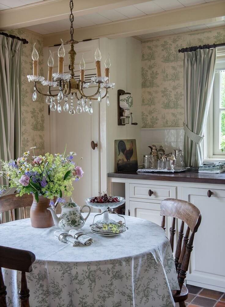 A rustic kitchen with an antique chandelier, a round kitchen table with a white and light green floral motif tablecloth, three brown vintage farmhouse chairs, a vase with spring flowers, a cake tray with cherries, silverware, and a vintage teapot.