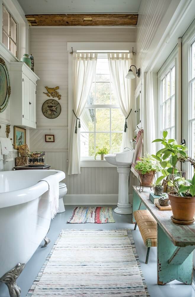 The interior of a vintage cottage bathroom with a white clawfoot bathtub, vintage rugs, a reclaimed turquoise-painted wooden bench with plants, two windows with cream-colored curtains, white paneled walls, a pedestal sink, and artwork on the walls.