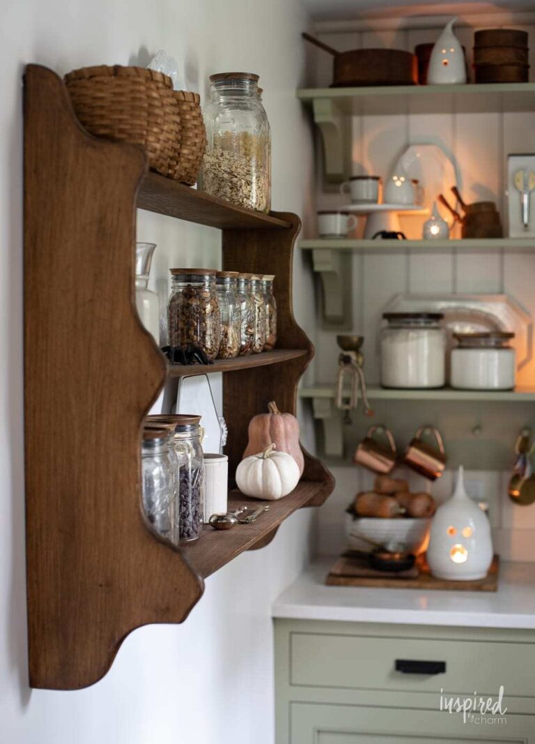 A dark brown antique kitchen cabinet shelf with spices in glass containers and more vintage kitchen shelves seen in the background, with copper mugs and other vintage accessories.