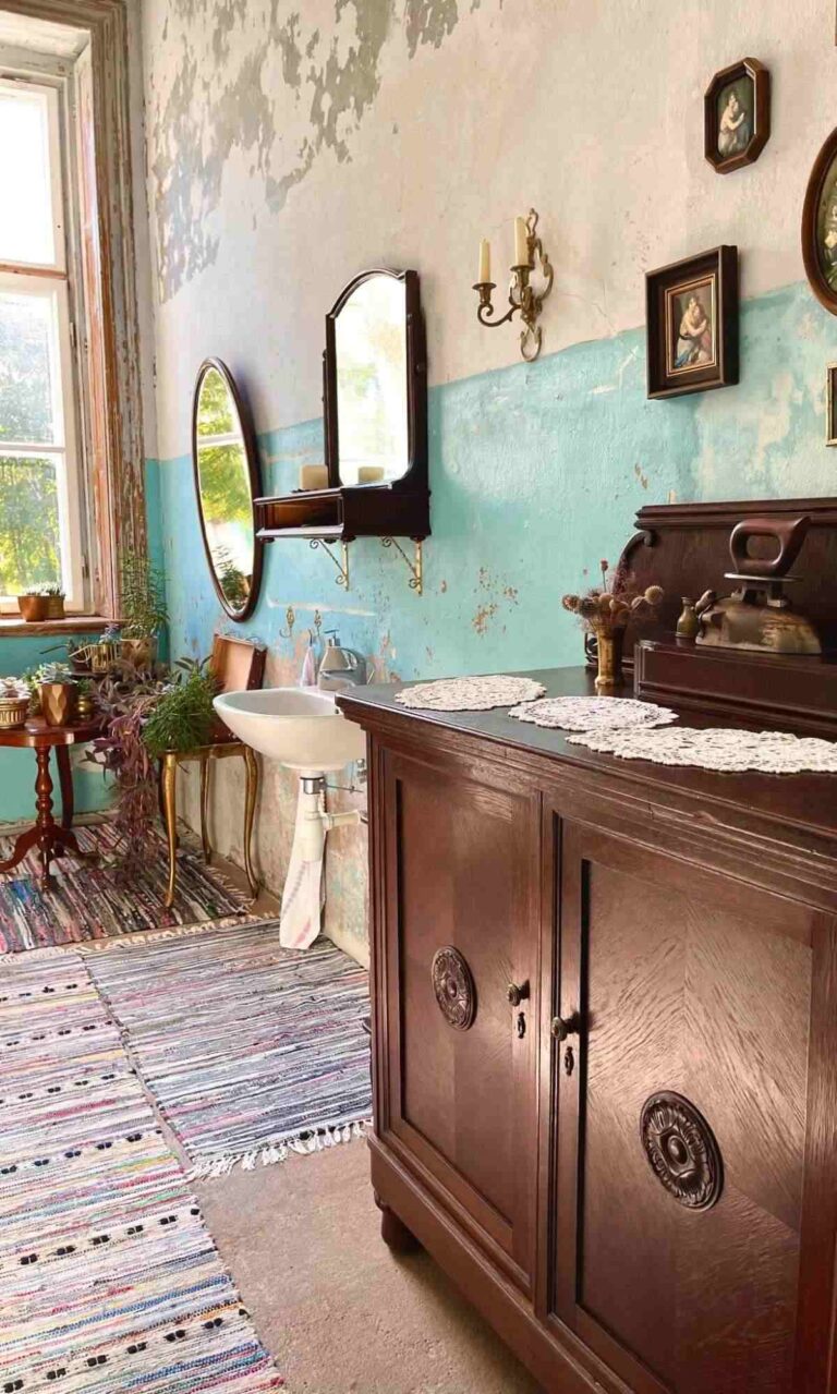 An interior of a vintage farmhouse bathroom featuring a dark wooden bathroom cabinet, a distressed white and turquoise wall, wall art, plants, a vintage sconce, and old-style mirrors.