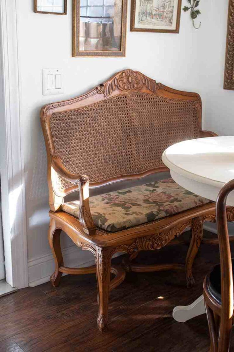 A decorative cane-back wooden bench with floral upholstery stands on a dark wood floor in a breakfast nook, with vintage frames hanging above it.