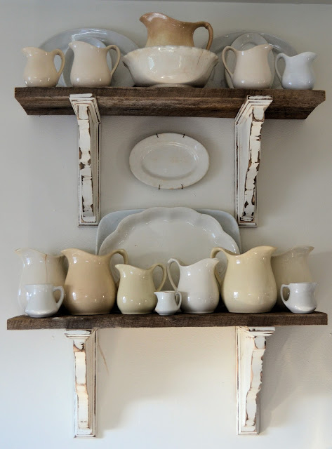 Two vintage shabby shelves hanging one above the other, with rustic white corbels and dark wood planks. Vintage white and cream milk jugs and other dishes stand on the shelves; a vintage white oval plate hangs between the two shelves.