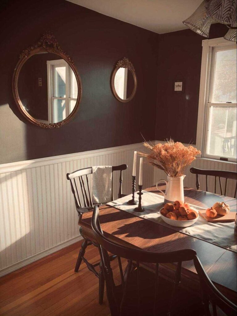 A view of a vintage wooden dining set with two candlesticks, a jug with a dried flower bouquet, and a plate of mini pumpkins. Two round vintage mirrors on a wainscoting wall