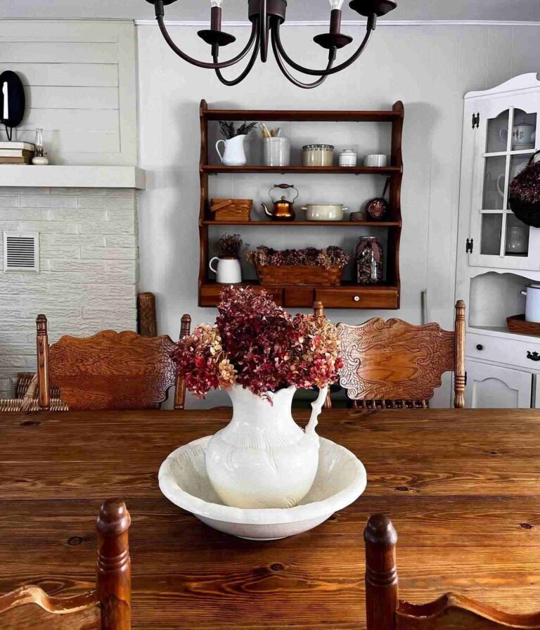 A view of a vintage farmhouse dining room with a wooden oak table and four vintage farmhouse chairs visible. A vintage dining room shelf cabinet with vintage dishes hangs on the wall in the background.