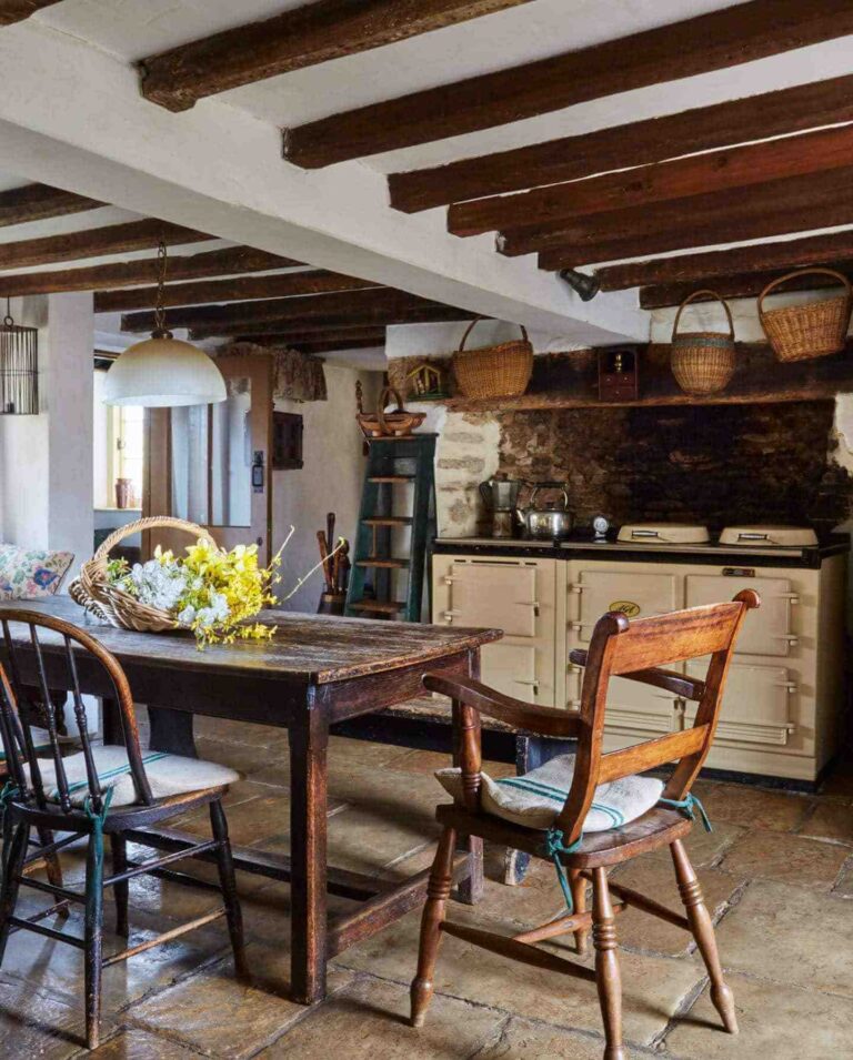 A dark wood table with mismatched vintage farmhouse chairs stands on a stone floor of a vintage farmhouse kitchen with dark wood beams and wicker baskets on the ceiling.