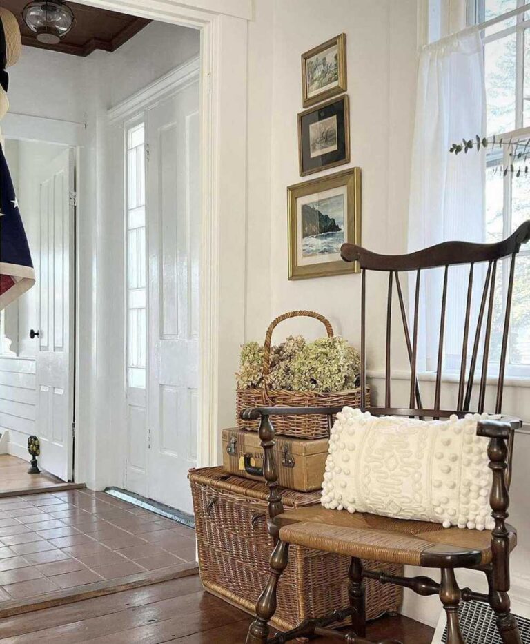 A coastal vintage farmhouse entryway with stone floor, a white door, a wicker storage trunk, a vintage suitcase, and a wicker basket with some dried flowers. A vintage dark wood hoop-back farmhouse chair stands on a wooden floor.