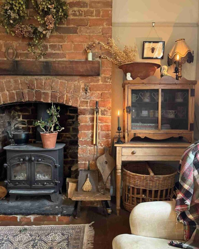 A cast-iron antique wood stove inside an old brick fireplace, with an old iron kettle and a plant on top. A vintage wooden cabinet stands on a vintage wood desk, decorated with a vintage table lamp and a vessel with dried flowers.