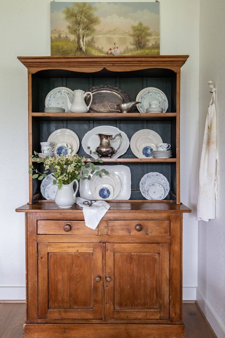 A vintage wooden hutch displaying antique and vintage dishes made of porcelain and pewter stands on a wooden floor and against a white wall. An old painting of a pastoral scene sits on top of the hutch.