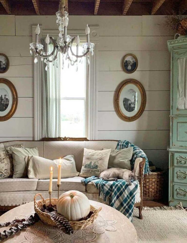 A vintage farmhouse decor interior with a vintage couch with vintage shabby-chic pillows, a cream-colored shiplap wall with oval gold-tone frames with old photos, a vintage turquoise cupboard, and a vintage chandelier.