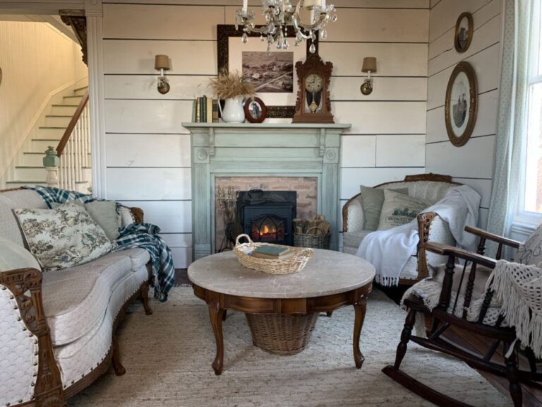 Vintage farmhouse living room with an antique wooden fireplace painted sage green and decorated with antique items such as an old clock, frame, and books. A vintage round coffee table stands in front of the fireplace, surrounded by an antique cream-colored couch, armchair, and a wooden rocking chair.